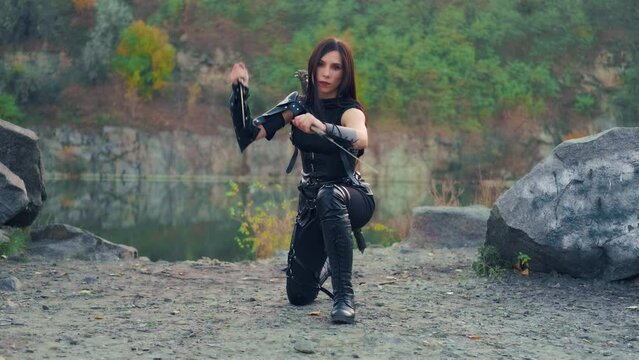Active fantasy woman warrior assassin holds with two blade daggers in hands spins waves with weapon. Lady dancer fighter. Armed sexy girl dancing. Black costume. Halloween concept. Green woods. 4k