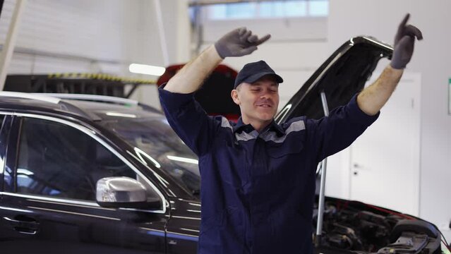 Smiling mechanic man in uniform and gloves dancing funny in a modern service auto center
