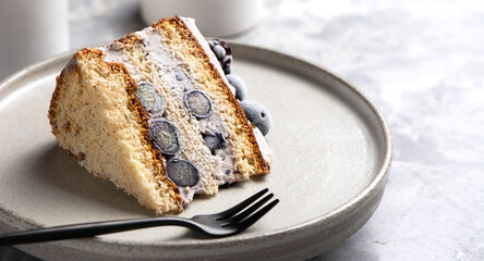 Slice of vanilla biscuit cake with blueberry cream and blackberries close-up on a ceramics plate on a light gray concrete background with copy space.
