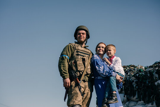 Woman with child boy on her hands stands near Ukrainian soldier on checkpoint against sandbags and sky background.