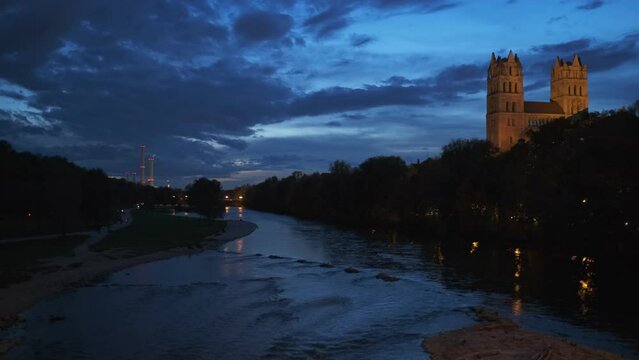 Munich nature at dramatic dusk: autumn in park of tourist Bavarian city. View of Isar river, trees and illuminated St Maximilian church from Reichenbach Bridge. Munchen, Bavaria, Germany.