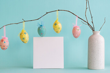 Colorful decorative Easter eggs hanging on dry branch placed in vase and blank card against blue background 