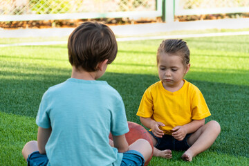 Upset little girl sitting barefoot near boy, talking with him and looking to ball while spending time together on green lawn during break from football game 