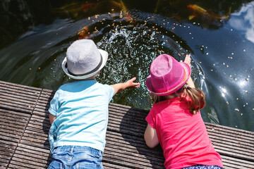2 kleine Kinder liegen auf einem Steg am Teich, planschen im Wasser und spielen mit Zierfischen
