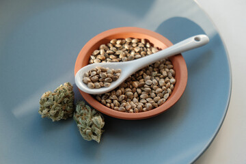 A bunch of cannabis seeds lie on a spoon and plate with several dried buds