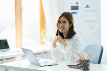 Obraz premium Portrait of Asian female executive sitting at office desk in front of laptop with financial business plan and smiling at camera at office