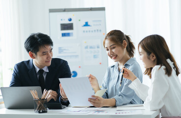 group of four happy young asian corporate people teammates meeting discussing business in office. Graphs on whiteboard behind them.