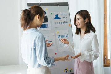 Fototapeta premium Two young businesswomen doing a presentation in a meeting or conference standing in front of a flip chart with hand drawn chart pointing to it with a pen