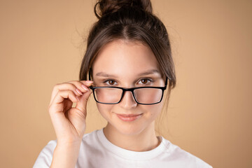 Young Caucasian girl in a white T-shirt takes off her glasses and smiles. Close-up portrait against a beige background.