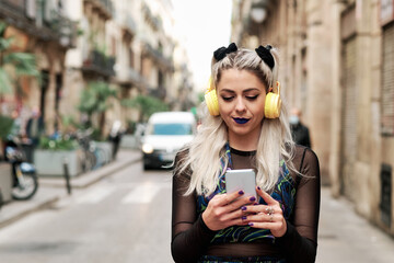 Obraz premium Young alternative woman using a mobile phone with headphones while walking down the street.