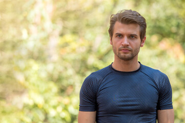 Portrait of attractive Caucasian athlete man in sports wear looking determined where he is going to with blurry background of trees.