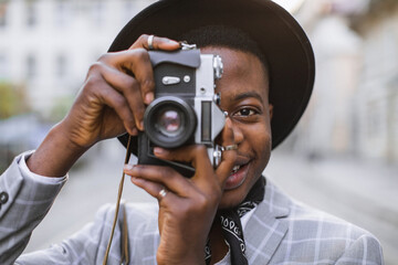 Smiling african guy in stylish suit and hat using vintage camera for taking photos outdoors. Handsome male hipster spending free time on fresh air.