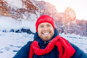 Fototapeta na wymiar Happy tourist man with ice on mustache background frozen Baikal lake in winter on sunny day in red hat