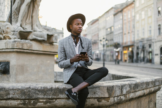 African american man in stylish suit and hat sitting on old fountain and holding modern smartphone. Handsome young guy surfing internet among city street.