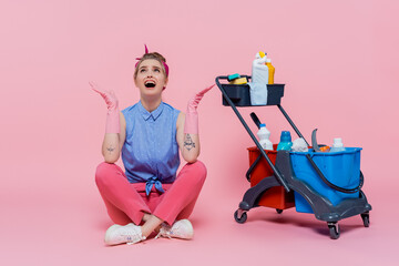 full length of crying young woman in rubber gloves sitting near housekeeping cart with cleaning supplies on pink.