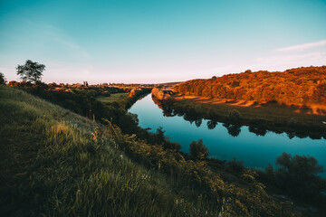 Malerische Landschaft in der Nähe der Stadt mit dem grünen Gras des Sonnenuntergangs und dem blauen Fluss