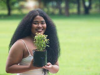 Beautiful dark skinned woman with long curly hair holding flowers in the black bag ready to plant in the park with copy space.