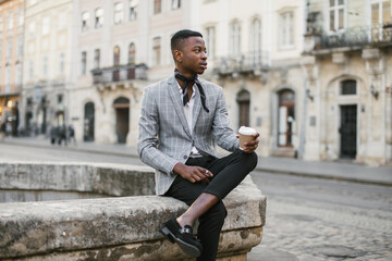 African american man in stylish suit sitting on old fountain with cup of coffee. Young male hipster...