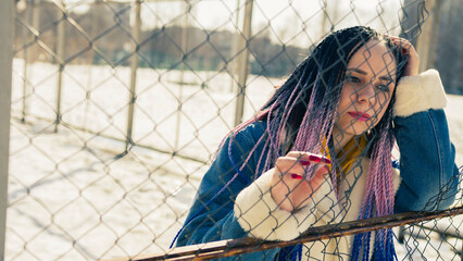 Fototapeta na wymiar Stylish woman leaning on metal fence. Serious young female in warm clothes looking away while leaning on net fence on city street