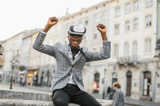 Overjoyed african guy in stylish suit emotionally gesturing on street while wearing VR goggles. Young businessman enjoying good news from work.