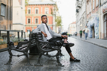 Confident african businessman in stylish suit sitting alone on bench among city street. Handsome...