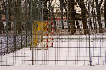 Boisko do piłki nożnej ( soccer ) , wewnątrz wysokiego ogrodzenia z siatki , zimą z leżącym śniegiem . Bramka piłkarska z siatką na tle drzew .