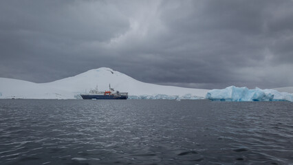 Ei Expeditionsschiff in den eisigen Gewässern des Südpolarmeeres vor einem riesigen Eisberg sowie weißen, schneebedeckten Gletschern