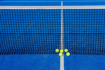 Five tennis balls next to a net of a blue paddle tennis court.
