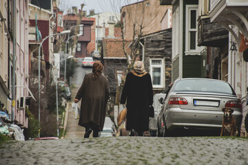 two neighborhood old women walking in street with head scarves and bags there is a dog looking in the corner