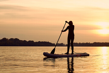 Silhouette of woman paddle on stand up paddle boarding (SUP) on quiet autumn river at twilight....