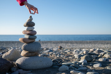 A hand puts a stone on a tower on the beach. Construction of a house by the sea. Background for...