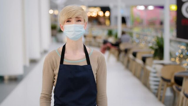 Friendly caucasian masked woman female waitress in medical face protective mask restaurant cafe kitchen bar worker looking at camera welcoming gesture come here approach welcome inviting invite client