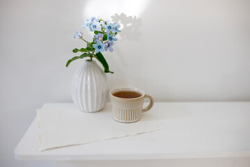 Fototapeta premium Flower hackelia velutina in a white fluted vase in the style of the seventies and a cup of coffee on the dresser. Scandinavian style