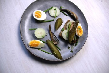 Plate with open sndwich with sprats, salty cucumber, onion, scallion, egg, bread on white background