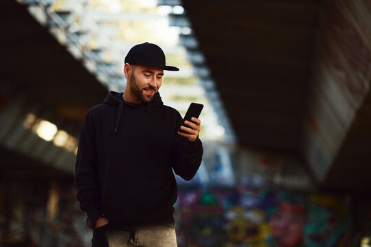 Young bearded man with a black hat  and black hoodie in the city using smartphone