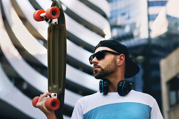 Young man with headphones around neck wearing hat and sunglasses holding a skateboard in his hand...