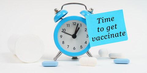 On a white surface, there are tablets and an alarm clock with a sticker that says - Time to get vaccinated
