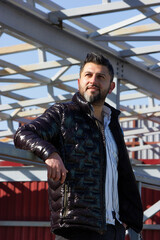 Smiling Turkish man is on the bridge, construction, looking somewhere in black jacket, cold season (autumn, winter, early spring), vertical plane