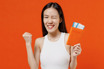 Young woman of Asian ethnicity 20s years old in white tank top hold passport boarding tickets doing winner gesture clenching fists say yes blinking isolated on plain orange background studio portrait.