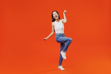 Fototapeta na wymiar Full size body length jubilant young woman of Asian ethnicity 20s year old in white tank top doing winner gesture celebrate clenching fists say yes isolated on plain orange background studio portrait
