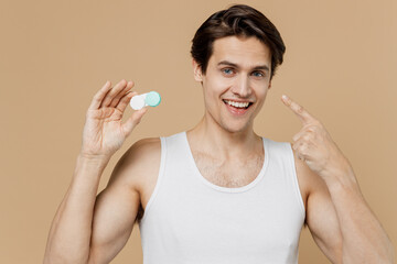 Attractive happy young man 20s perfect skin in undershirt hold contact lenses case point finger on container isolated on pastel pastel beige background studio Eyecare healthcare procedures concept