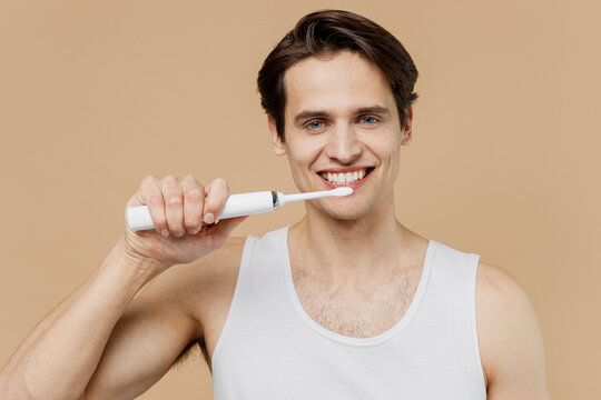 Attractive cool young man 20s perfect skin wearing undershirt hold brush brushing teeth isolated on pastel pastel beige background studio portrait. Skin care healthcare cosmetic procedures concept