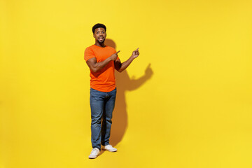 Full body young man of African American ethnicity 20s in orange t-shirt point index finger aside on workspace area mock up isolated on plain yellow background studio portrait People lifestyle concept