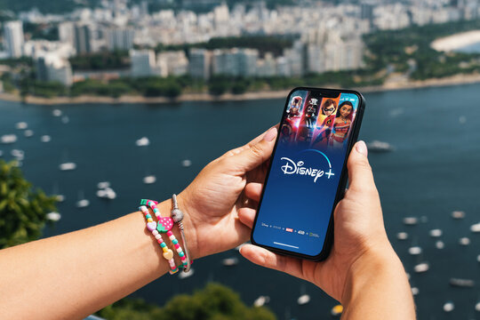 Girl holding smartphone with Disney+ (Disney Plus) app on screen. City and bay with some boats in the background. Rio de Janeiro, RJ, Brazil. March 2022