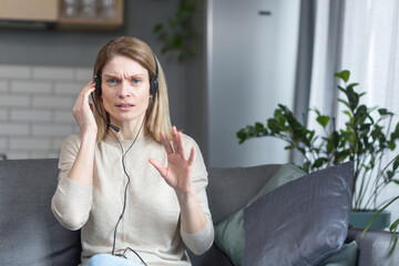 Angry woman talking on video call looks at camera and explains uses headset, works remotely from...