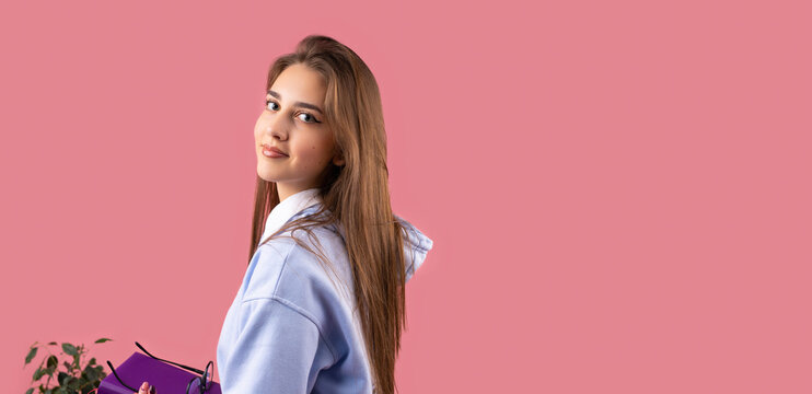 Picture of a young beautiful smiling student girl with blond long hair and make-up looking at the camera over her shoulder on pink background and copy space.