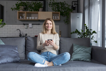 Fototapeta premium Beautiful middle-aged woman sitting on the couch at home, using the phone and smiling