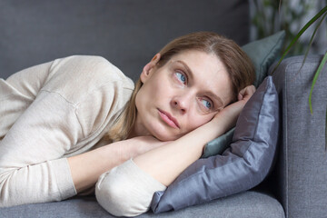 Close-up photo of a pensive middle-aged woman lying on a sofa, looking out the window at home