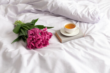 Fototapeta na wymiar Bouquet of pink tulips, cup of tea and book on bed with lilac linen.