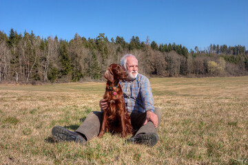 A man is sitting in a meadow. His Irish Setter dog is sitting between his legs. Experiencing nature...